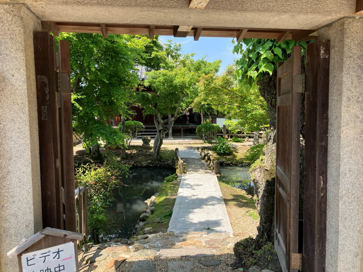 新薬師寺の庭への入口 — 門の先に池と石橋、ビデオ上映中の案内板