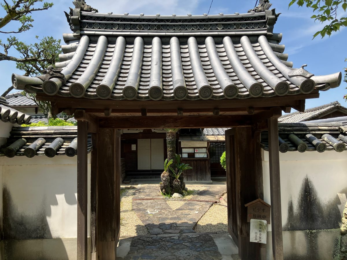 新薬師寺の庭へのもうひとつの入口 — 瓦屋根の門と「ビデオ放映中」「七佛薬師金堂跡 出土品」の札