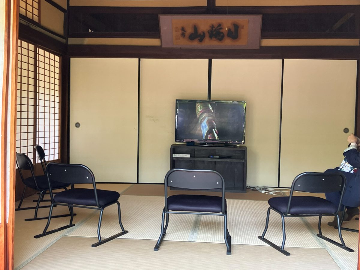 新薬師寺のビデオ上映の部屋 — 畳の間に椅子が並び、山号「日輪山」の額がかかる